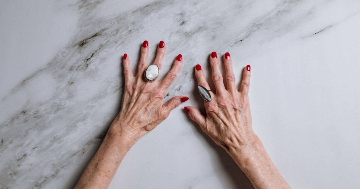Two hands with red nails resting on a grey-veined marble surface, the stone pattern that marble nail photos have to reproduce on the nail