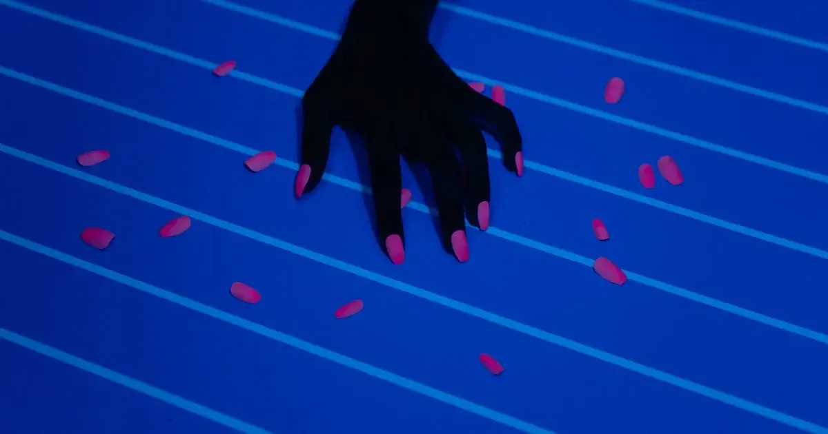 A hand with a brightly coloured manicure surrounded by nail polish bottles, the kind of neon set that clips in phone photos