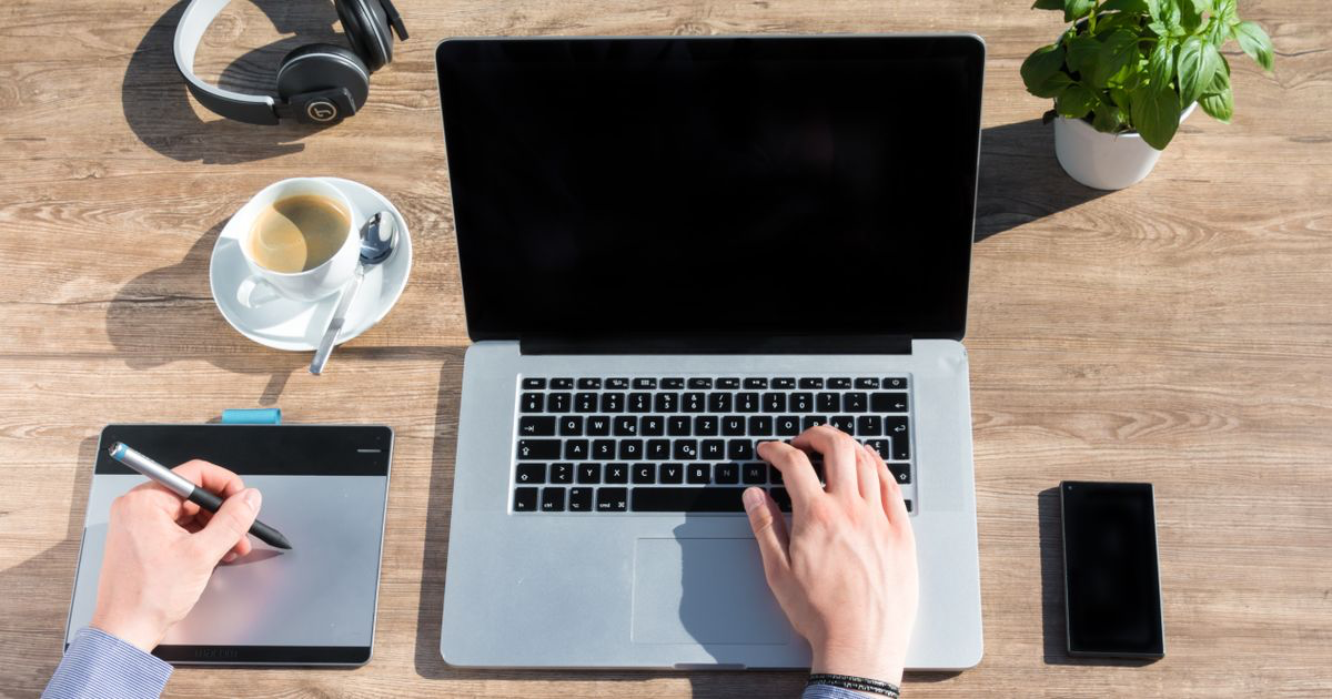 A laptop, drawing tablet, phone and coffee laid out on a wooden desk, the kind of setup used for making nail salon graphics in Canva