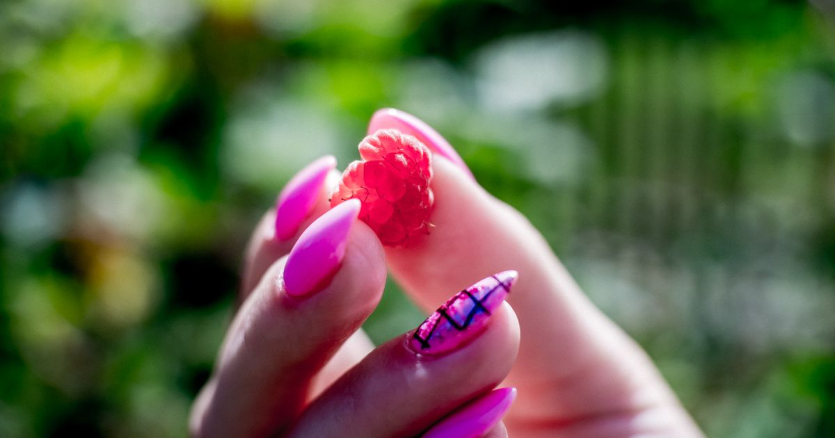 A hand with sheer pink nails holding a raspberry, the kind of translucent finish that needs careful editing on jelly nail photos