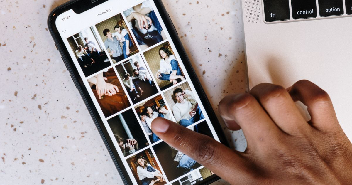Close-up of a person browsing a social media app on a smartphone beside a laptop, reviewing nail photos for a portfolio