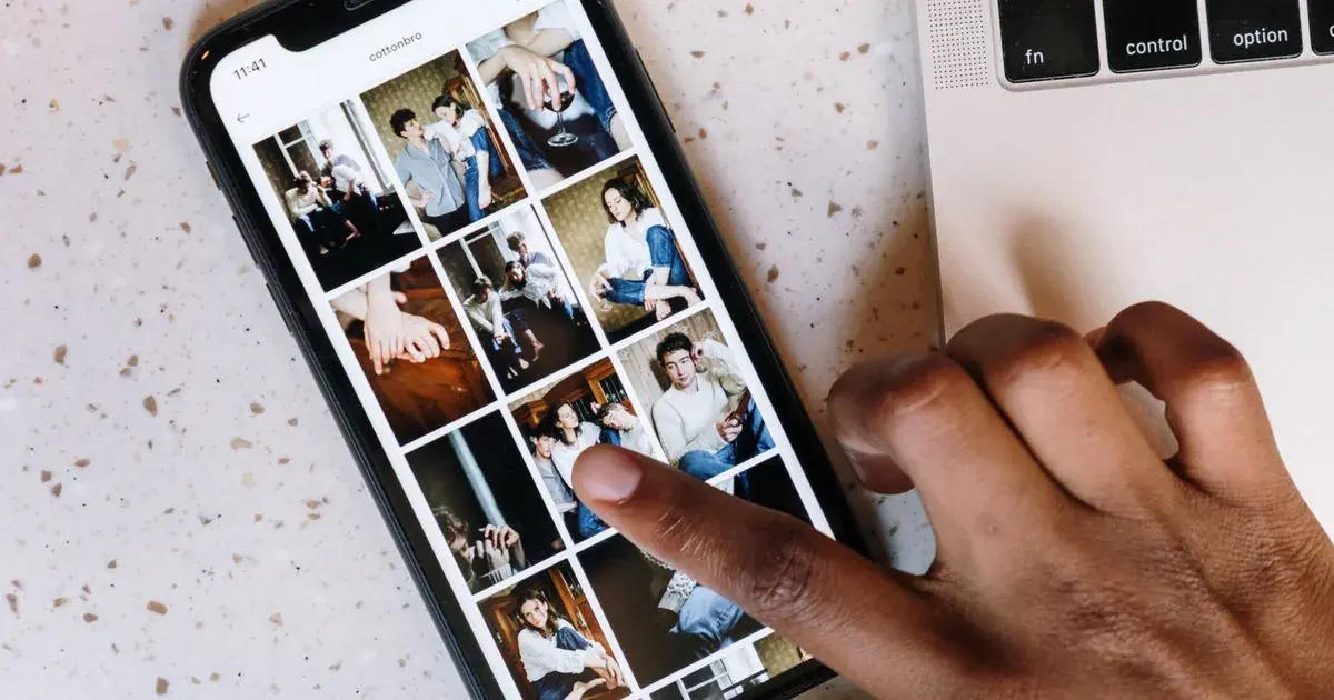 Close-up of a person browsing a social media app on a smartphone beside a laptop, reviewing nail photos for a portfolio