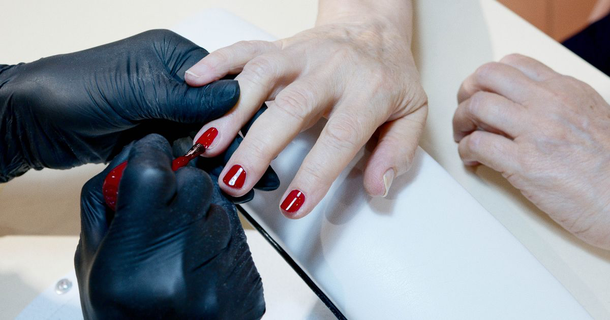 A manicurist applying deep red polish to a client's nails in a salon, the kind of jewel tone used for velvet nail photos