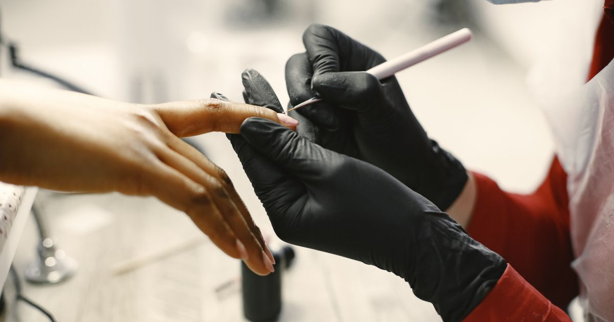 A manicurist in black gloves cleaning a client's fingernails at a salon desk