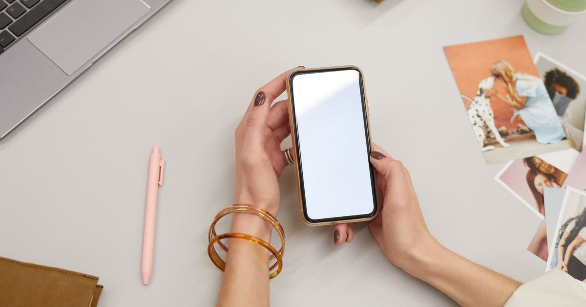 A person holding a smartphone at a desk beside a laptop and printed photos, planning a nail portfolio website