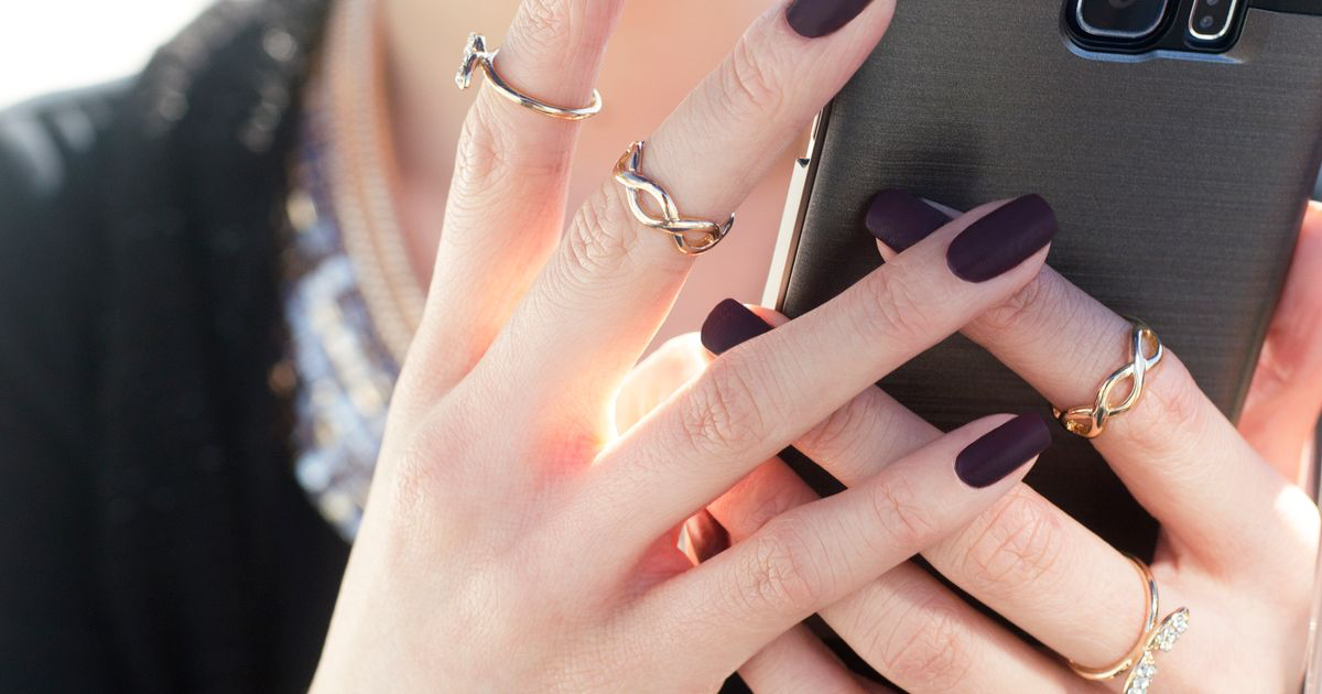 Woman with manicured nails and gold rings holding a smartphone, considering VSCO for nail photos